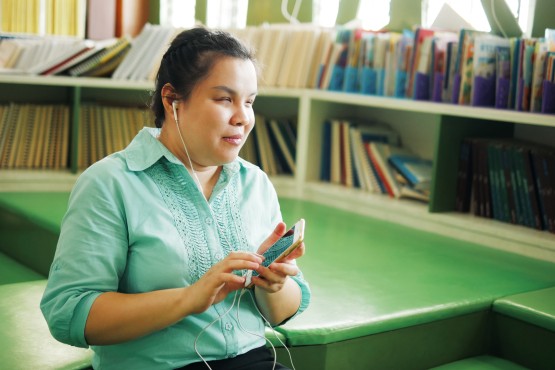 A Blind Asian woman is wearing earphones using a smart phone with voice accessibility in a library.